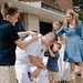 A U.S. Navy officer in white dress uniform is being embraced and pinned by his children during what appears to be a military promotion or retirement ceremony. The scene takes place outdoors in front of a brick building, with a United States Marine Corps flag visible in the background. The moment is joyful and celebratory, with multiple children participating in the pinning tradition.