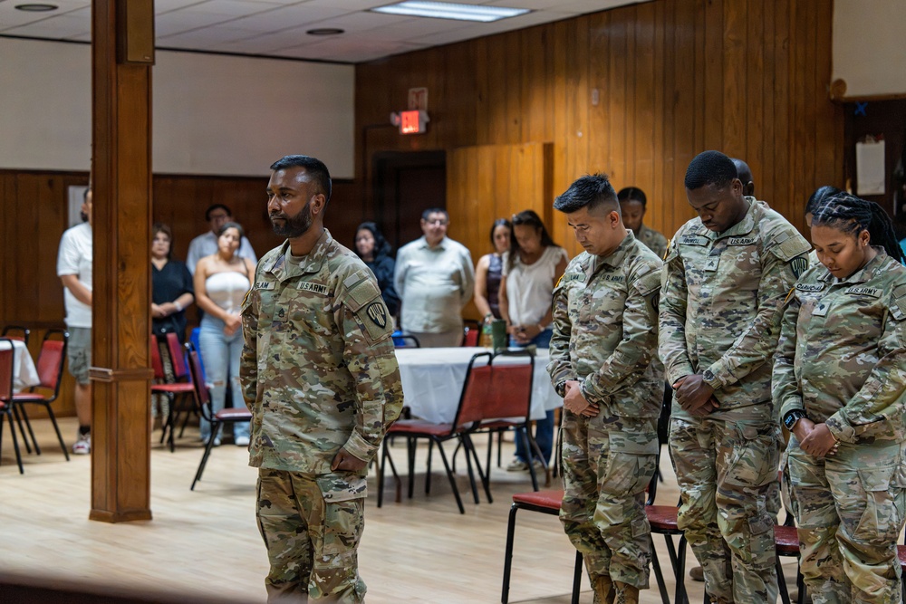 Four U.S. Army soldiers in OCP (Operational Camouflage Pattern) uniforms stand in a formal indoor setting, with three bowing their heads in what appears to be a moment of prayer or reflection. Civilians stand in the background observing the ceremony. The setting appears to be a community hall or event space with wood-paneled walls.