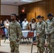 Four U.S. Army soldiers in OCP (Operational Camouflage Pattern) uniforms stand in a formal indoor setting, with three bowing their heads in what appears to be a moment of prayer or reflection. Civilians stand in the background observing the ceremony. The setting appears to be a community hall or event space with wood-paneled walls.