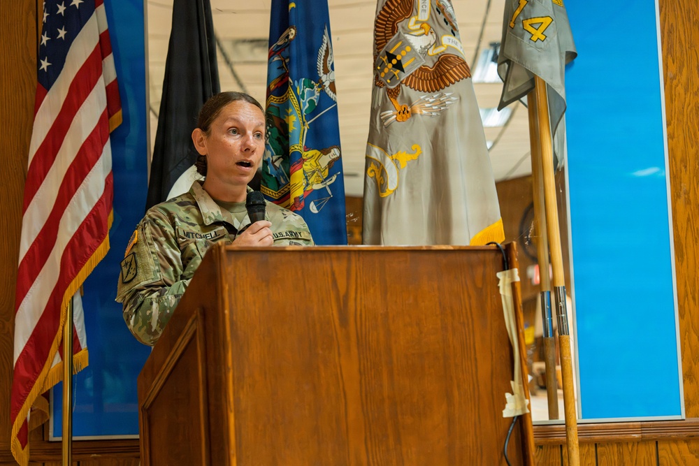 A U.S. Army soldier, identified by name tape as 'Mitchell,' is speaking at a wooden podium while holding a microphone. The setting appears to be an official military ceremony or briefing, with multiple flags displayed in the background. The soldier is wearing Army Combat Uniform (ACU) with a U.S. Army chest tape visible.