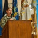 A U.S. Army soldier, identified by name tape as 'Mitchell,' is speaking at a wooden podium while holding a microphone. The setting appears to be an official military ceremony or briefing, with multiple flags displayed in the background. The soldier is wearing Army Combat Uniform (ACU) with a U.S. Army chest tape visible.