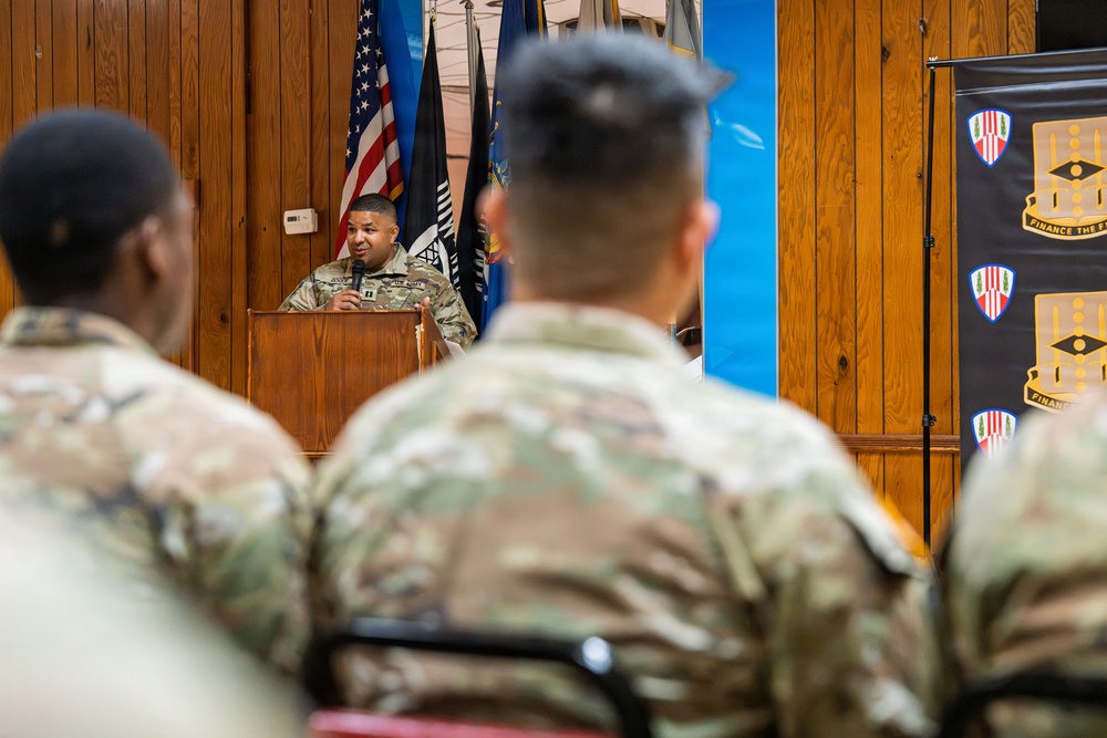 A U.S. Army officer in OCP (Operational Camouflage Pattern) uniform is speaking at a wooden podium during a military ceremony or briefing. An audience of soldiers in matching uniforms is seated facing the speaker. A banner on the right reads 'Finance the F...' suggesting this is a Finance Corps unit event.