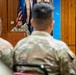 A U.S. Army officer in OCP (Operational Camouflage Pattern) uniform is speaking at a wooden podium during a military ceremony or briefing. An audience of soldiers in matching uniforms is seated facing the speaker. A banner on the right reads 'Finance the F...' suggesting this is a Finance Corps unit event.