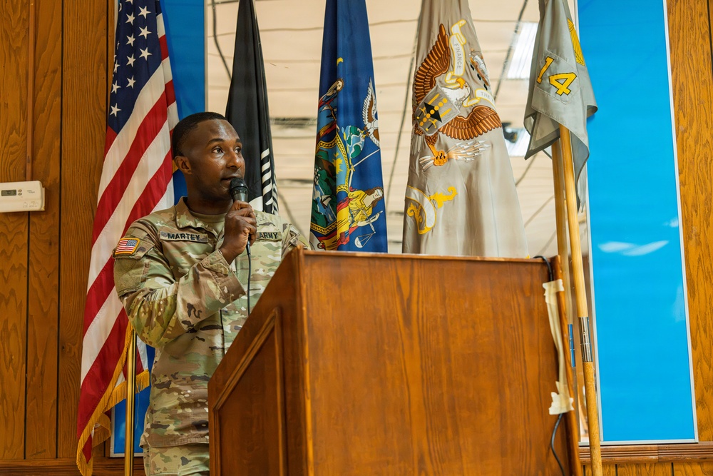A U.S. Army soldier named Martey is speaking at a wooden podium while holding a microphone. The setting appears to be an official military ceremony or briefing, with multiple flags displayed in the background including the U.S. flag, POW/MIA flag, a state flag (likely New York), and military unit flags. The soldier is wearing Army Combat Uniform (ACU) in OCP (Operational Camouflage Pattern).