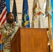 A U.S. Army soldier named Martey is speaking at a wooden podium while holding a microphone. The setting appears to be an official military ceremony or briefing, with multiple flags displayed in the background including the U.S. flag, POW/MIA flag, a state flag (likely New York), and military unit flags. The soldier is wearing Army Combat Uniform (ACU) in OCP (Operational Camouflage Pattern).