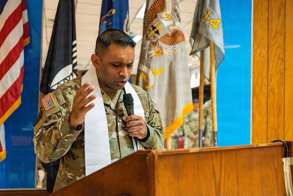 A U.S. Army chaplain in OCP (Operational Camouflage Pattern) uniform is delivering a prayer or invocation at a military ceremony. He is wearing a white liturgical stole over his uniform, holding a microphone, and standing at a wooden podium. Multiple military flags and colors are displayed in the background.