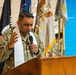 A U.S. Army chaplain in OCP (Operational Camouflage Pattern) uniform is delivering a prayer or invocation at a military ceremony. He is wearing a white liturgical stole over his uniform, holding a microphone, and standing at a wooden podium. Multiple military flags and colors are displayed in the background.