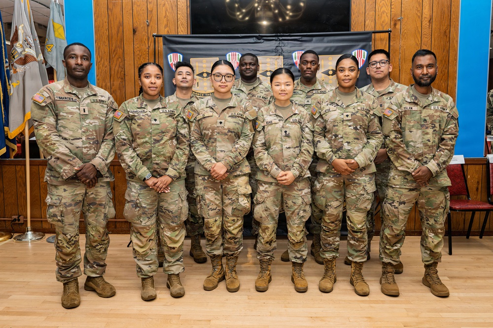 A group of approximately eleven U.S. Army soldiers in Operational Camouflage Pattern (OCP) uniforms pose for a formal group photo indoors. They are standing in two rows in front of a backdrop displaying military unit insignia/patches. The setting appears to be a military community or event hall with wooden paneling and flags visible.