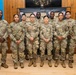 A group of approximately eleven U.S. Army soldiers in Operational Camouflage Pattern (OCP) uniforms pose for a formal group photo indoors. They are standing in two rows in front of a backdrop displaying military unit insignia/patches. The setting appears to be a military community or event hall with wooden paneling and flags visible.