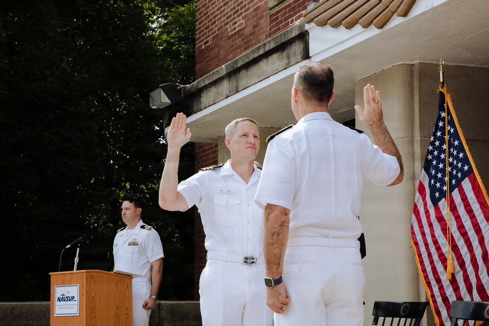 A formal U.S. Navy military ceremony is taking place outdoors, likely a promotion or re-enlistment oath ceremony. Two naval officers in white dress uniforms face each other with right hands raised, administering or receiving an oath. A third officer stands at a NAVSUP-branded podium in the background.