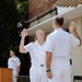 A formal U.S. Navy military ceremony is taking place outdoors, likely a promotion or re-enlistment oath ceremony. Two naval officers in white dress uniforms face each other with right hands raised, administering or receiving an oath. A third officer stands at a NAVSUP-branded podium in the background.