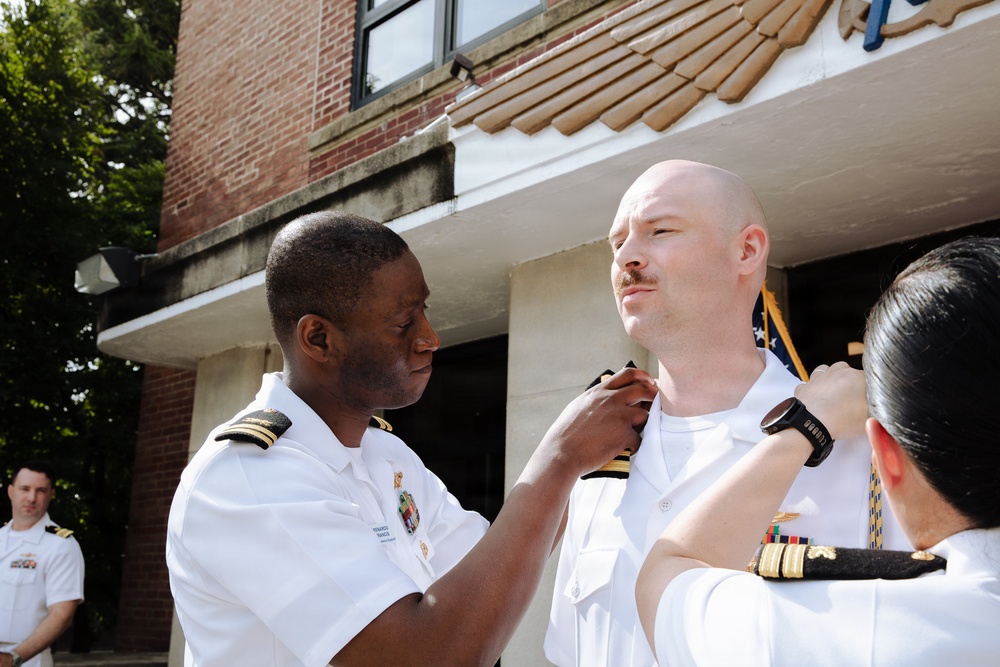 A military promotion or pinning ceremony is taking place outdoors in front of a brick building. Two individuals in white naval uniforms are simultaneously pinning insignia or rank devices onto the collar/shoulder area of a bald man with a mustache in a white naval uniform. A fourth uniformed officer stands in the background observing the ceremony.