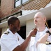 A military promotion or pinning ceremony is taking place outdoors in front of a brick building. Two individuals in white naval uniforms are simultaneously pinning insignia or rank devices onto the collar/shoulder area of a bald man with a mustache in a white naval uniform. A fourth uniformed officer stands in the background observing the ceremony.