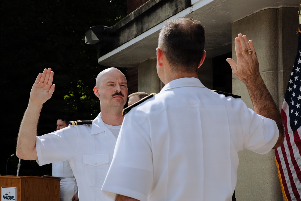 A military swearing-in ceremony is taking place outdoors, with two naval officers in white dress uniforms raising their right hands. One officer faces the camera while administering or receiving an oath, with an American flag visible in the background. A podium bearing a 'NAVSUP' placard is visible to the left.
