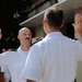 A military swearing-in ceremony is taking place outdoors, with two naval officers in white dress uniforms raising their right hands. One officer faces the camera while administering or receiving an oath, with an American flag visible in the background. A podium bearing a 'NAVSUP' placard is visible to the left.