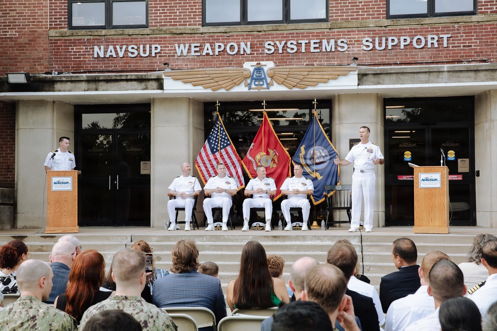 A formal military ceremony is taking place outside the NAVSUP Weapon Systems Support building. Several naval officers in white dress uniforms are on a raised platform, with one speaking at a podium while four others are seated. An audience of civilians and military personnel is gathered in front of the steps.
