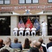 A formal military ceremony is taking place outside the NAVSUP Weapon Systems Support building. Several naval officers in white dress uniforms are on a raised platform, with one speaking at a podium while four others are seated. An audience of civilians and military personnel is gathered in front of the steps.