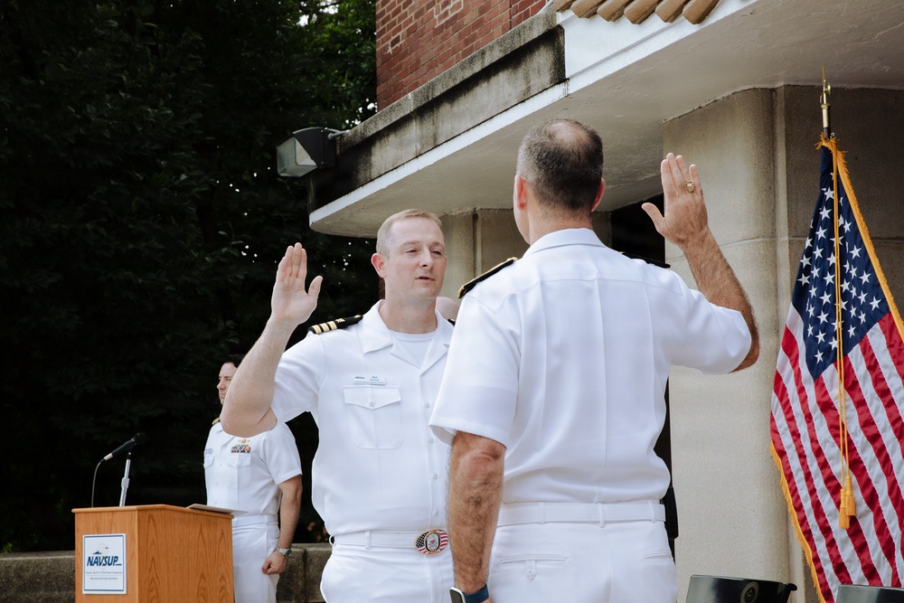 A U.S. Navy military ceremony is taking place outdoors, appearing to be a swearing-in or promotion ceremony. Two naval officers in white dress uniforms face each other with right hands raised, suggesting an oath is being administered. A podium bearing the NAVSUP insignia is visible in the background alongside an American flag with gold fringe.