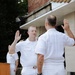 A U.S. Navy military ceremony is taking place outdoors, appearing to be a swearing-in or promotion ceremony. Two naval officers in white dress uniforms face each other with right hands raised, suggesting an oath is being administered. A podium bearing the NAVSUP insignia is visible in the background alongside an American flag with gold fringe.
