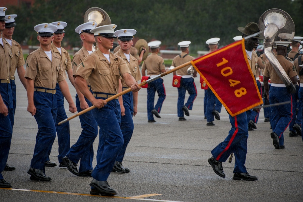 A U.S. Marine Corps graduation or parade ceremony is taking place on a parade deck in wet, rainy conditions. Marines in service uniforms (khaki blouses, blue trousers, white covers) are marching in formation, with a guidon bearer carrying a red pennant marked '1048'. A military band with brass instruments, including sousaphones, is visible in the background.