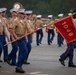 A U.S. Marine Corps graduation or parade ceremony is taking place on a parade deck in wet, rainy conditions. Marines in service uniforms (khaki blouses, blue trousers, white covers) are marching in formation, with a guidon bearer carrying a red pennant marked '1048'. A military band with brass instruments, including sousaphones, is visible in the background.