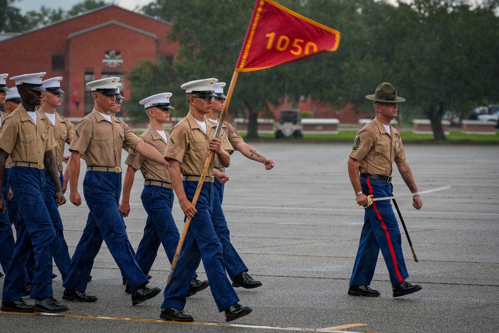 A formation of U.S. Marines in service uniforms marches across a parade deck, led by a Drill Instructor wearing a distinctive campaign cover (Smokey Bear hat). One Marine carries a red guidon flag marked '1050', indicating the unit or platoon designation. The scene appears to be a graduation ceremony or formal drill event at a Marine Corps base.