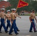 A formation of U.S. Marines in service uniforms marches across a parade deck, led by a Drill Instructor wearing a distinctive campaign cover (Smokey Bear hat). One Marine carries a red guidon flag marked '1050', indicating the unit or platoon designation. The scene appears to be a graduation ceremony or formal drill event at a Marine Corps base.