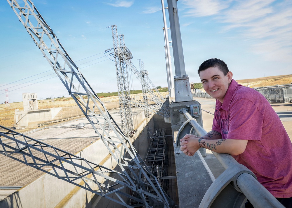 A young person wearing a pink/magenta polo shirt leans against a concrete railing at what appears to be a dam or water control facility. They are smiling at the camera with visible tattoos on their forearm. The background features multiple high-voltage electrical transmission towers and concrete infrastructure under a partly cloudy sky.