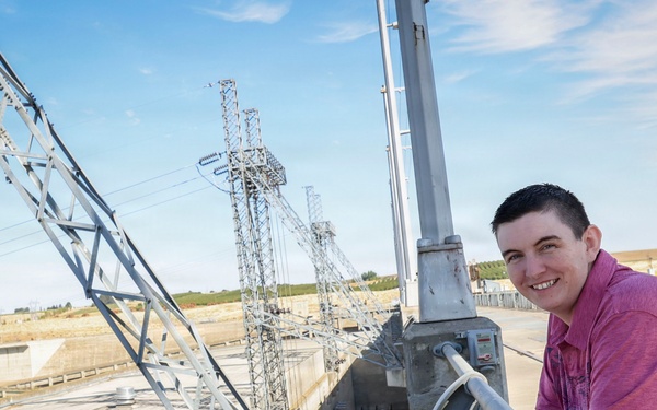 A young person wearing a pink/magenta polo shirt leans against a concrete railing at what appears to be a dam or water control facility. They are smiling at the camera with visible tattoos on their forearm. The background features multiple high-voltage electrical transmission towers and concrete infrastructure under a partly cloudy sky.