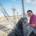 A young person wearing a pink/magenta polo shirt leans against a concrete railing at what appears to be a dam or water control facility. They are smiling at the camera with visible tattoos on their forearm. The background features multiple high-voltage electrical transmission towers and concrete infrastructure under a partly cloudy sky.