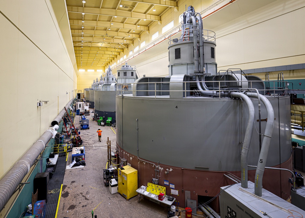 The image shows the interior of a large industrial facility, likely a hydroelectric power plant or similar power generation station, featuring a row of large cylindrical turbine/generator units numbered sequentially along a wide service corridor. The facility is well-lit with a high ceiling and yellow-painted walls, and appears to be undergoing maintenance or refurbishment work. Multiple pieces of equipment, tools, and work stations are visible along the floor between the machinery and the wall.