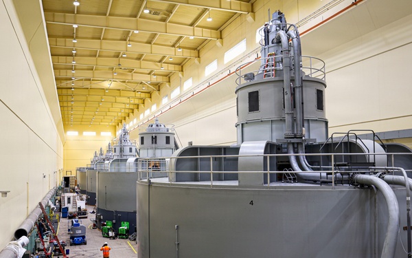 The image shows the interior of a large industrial facility, likely a hydroelectric power plant or similar power generation station, featuring a row of large cylindrical turbine/generator units numbered sequentially along a wide service corridor. The facility is well-lit with a high ceiling and yellow-painted walls, and appears to be undergoing maintenance or refurbishment work. Multiple pieces of equipment, tools, and work stations are visible along the floor between the machinery and the wall.