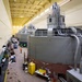 The image shows the interior of a large industrial facility, likely a hydroelectric power plant or similar power generation station, featuring a row of large cylindrical turbine/generator units numbered sequentially along a wide service corridor. The facility is well-lit with a high ceiling and yellow-painted walls, and appears to be undergoing maintenance or refurbishment work. Multiple pieces of equipment, tools, and work stations are visible along the floor between the machinery and the wall.
