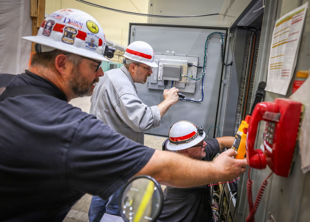 Three workers wearing white hard hats with red stripes are performing electrical or telecommunications maintenance work on an industrial control panel or equipment cabinet. The workers appear to be technicians or electricians collaborating on installation or troubleshooting tasks. Various tools and equipment are visible, including a multimeter and a red emergency telephone mounted on the cabinet.