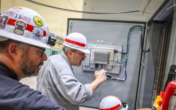 Three workers wearing white hard hats with red stripes are performing electrical or telecommunications maintenance work on an industrial control panel or equipment cabinet. The workers appear to be technicians or electricians collaborating on installation or troubleshooting tasks. Various tools and equipment are visible, including a multimeter and a red emergency telephone mounted on the cabinet.