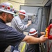 Three workers wearing white hard hats with red stripes are performing electrical or telecommunications maintenance work on an industrial control panel or equipment cabinet. The workers appear to be technicians or electricians collaborating on installation or troubleshooting tasks. Various tools and equipment are visible, including a multimeter and a red emergency telephone mounted on the cabinet.