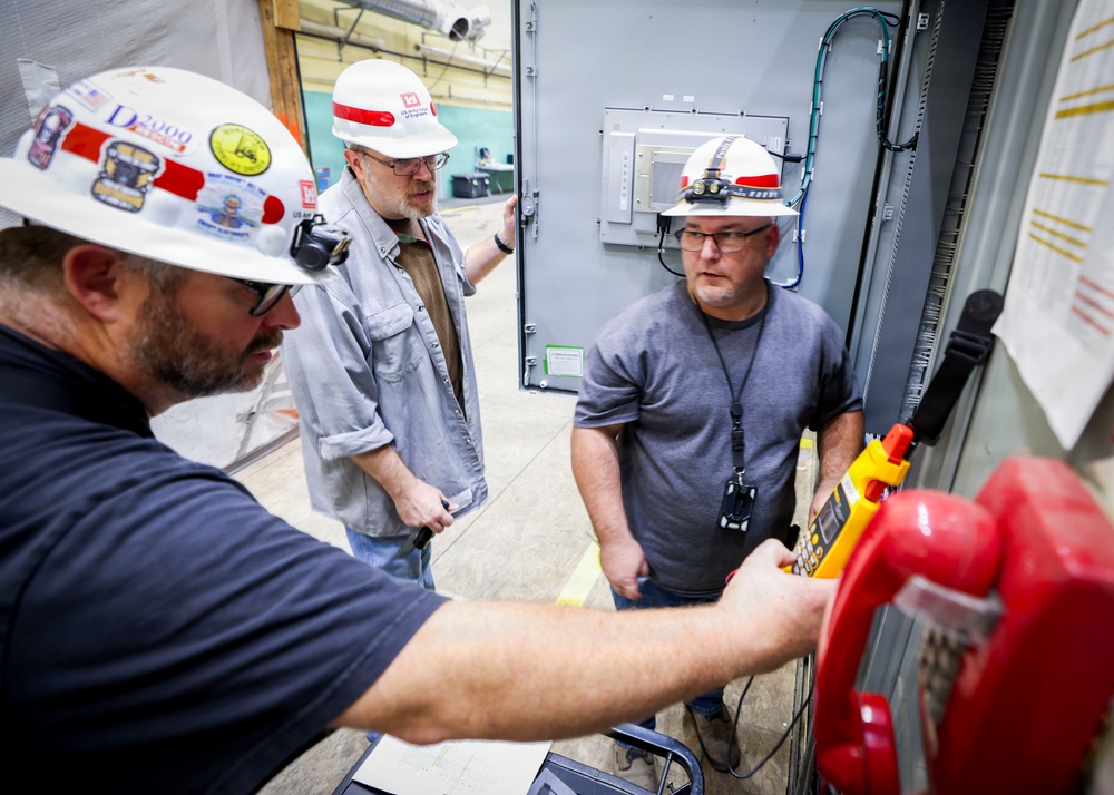 Three workers wearing white hard hats with red stripes are gathered around an electrical control panel or equipment cabinet in an industrial facility. One worker in the foreground is operating a yellow and red handheld control device, while the other two observe closely. The setting appears to be an active industrial or construction site with electrical infrastructure visible.