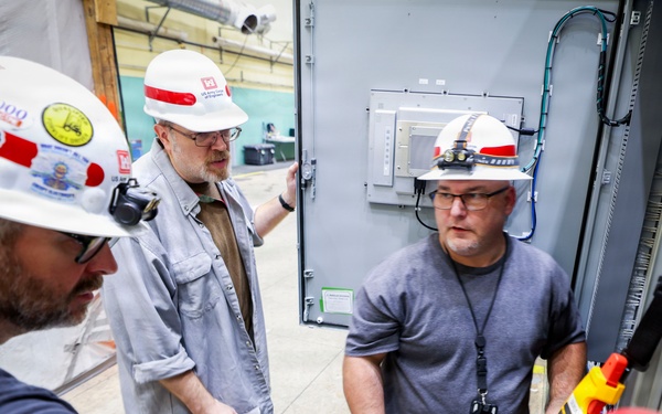 Three workers wearing white hard hats with red stripes are gathered around an electrical control panel or equipment cabinet in an industrial facility. One worker in the foreground is operating a yellow and red handheld control device, while the other two observe closely. The setting appears to be an active industrial or construction site with electrical infrastructure visible.
