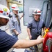 Three workers wearing white hard hats with red stripes are gathered around an electrical control panel or equipment cabinet in an industrial facility. One worker in the foreground is operating a yellow and red handheld control device, while the other two observe closely. The setting appears to be an active industrial or construction site with electrical infrastructure visible.