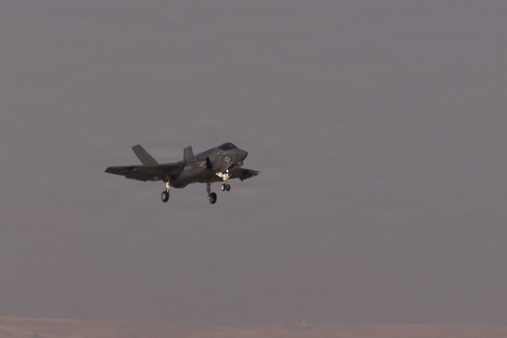 A fifth-generation stealth fighter jet is captured in flight with its landing gear fully deployed, suggesting it is on approach for landing. The aircraft displays a dark gray low-visibility paint scheme consistent with modern military stealth aircraft. The background shows an overcast, hazy sky with a faint horizon line visible at the bottom of the frame.