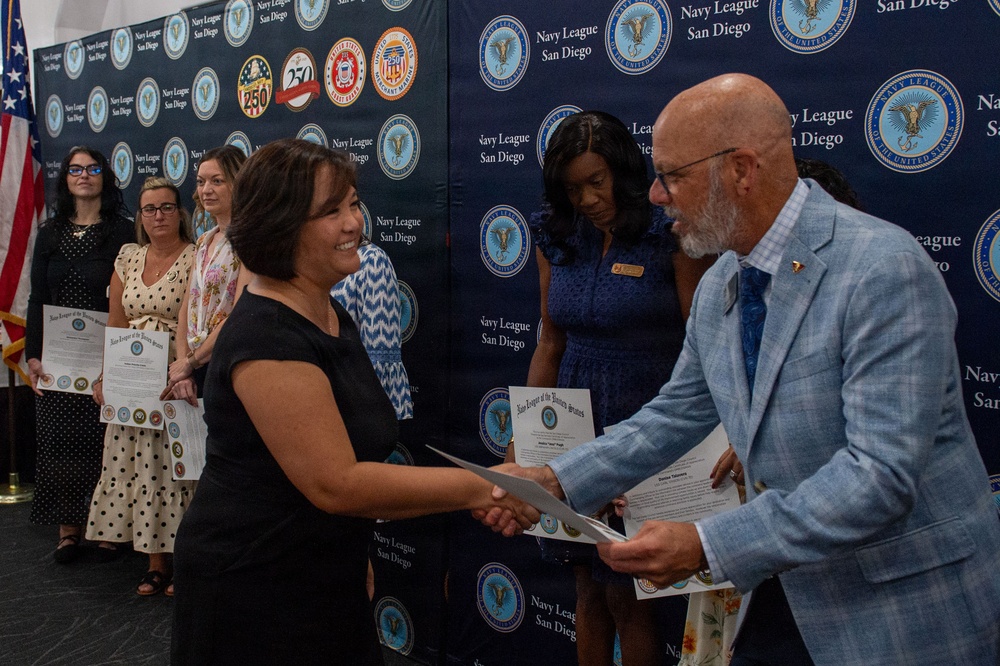 A formal award or recognition ceremony hosted by the Navy League San Diego, where a man in a light blue blazer is presenting a certificate to a smiling woman in a black dress. Several other women stand in the background, also holding certificates, suggesting multiple recipients are being honored. The event takes place in front of a branded step-and-repeat backdrop featuring Navy League San Diego logos and military insignia.