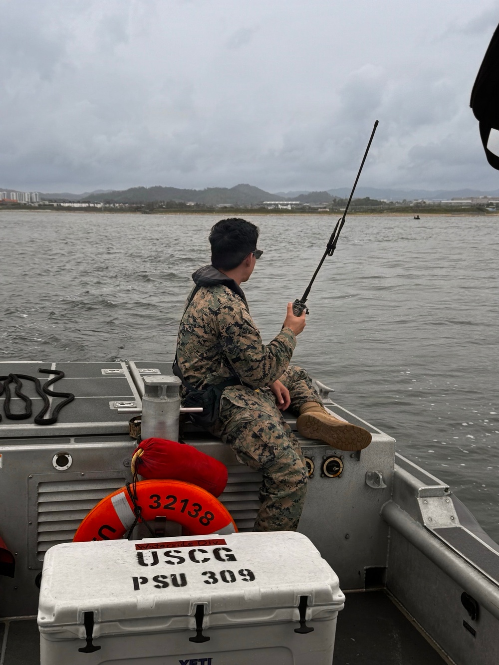 A person in military camouflage (MARPAT pattern) sits on the gunwale of a USCG PSU 309 vessel, holding a handheld radio with an extended antenna. The boat is on open water with a coastal cityscape and hills visible in the background under overcast skies. Equipment including an orange life ring marked '32138' and a YETI cooler stenciled 'USCG PSU 309' are visible in the foreground.