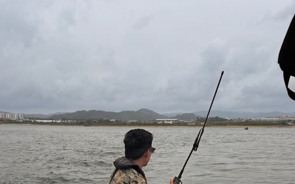 A person in military camouflage (MARPAT pattern) sits on the gunwale of a USCG PSU 309 vessel, holding a handheld radio with an extended antenna. The boat is on open water with a coastal cityscape and hills visible in the background under overcast skies. Equipment including an orange life ring marked '32138' and a YETI cooler stenciled 'USCG PSU 309' are visible in the foreground.