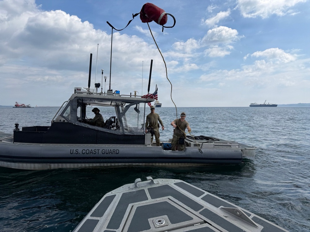 A U.S. Coast Guard patrol boat is operating on open water with three personnel visible on deck. One crew member is throwing or catching a line with a red buoy attached, suggesting a line-transfer or mooring operation between two vessels. Several large ships are visible in the background on the horizon.