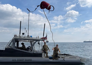 A U.S. Coast Guard patrol boat is operating on open water with three personnel visible on deck. One crew member is throwing or catching a line with a red buoy attached, suggesting a line-transfer or mooring operation between two vessels. Several large ships are visible in the background on the horizon.