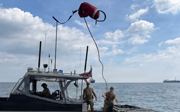 A U.S. Coast Guard patrol boat is operating on open water with three personnel visible on deck. One crew member is throwing or catching a line with a red buoy attached, suggesting a line-transfer or mooring operation between two vessels. Several large ships are visible in the background on the horizon.