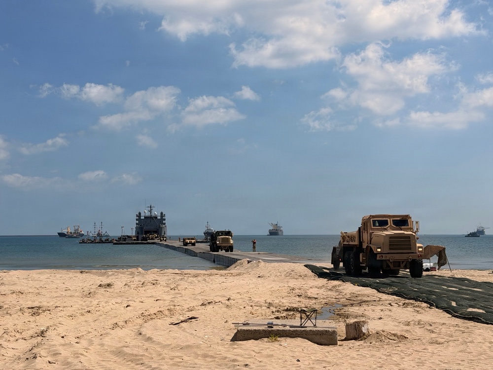 A military amphibious logistics operation is underway on a sandy beach, featuring a causeway or floating pier extending from the shore to a vessel offshore. Military trucks in tan/desert camouflage are staged on the beach and moving along the pier. Multiple vessels are visible offshore in calm waters under partly cloudy skies.