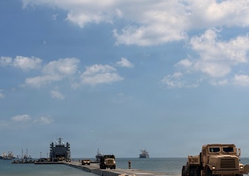 A military amphibious logistics operation is underway on a sandy beach, featuring a causeway or floating pier extending from the shore to a vessel offshore. Military trucks in tan/desert camouflage are staged on the beach and moving along the pier. Multiple vessels are visible offshore in calm waters under partly cloudy skies.