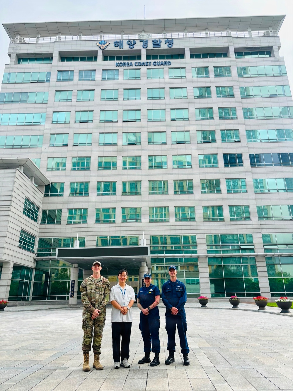 Four individuals are posing for a group photo in front of the Korea Coast Guard (해양경찰청) headquarters building. The group includes one person in U.S. Army multicam/OCP camouflage uniform, one civilian in a white shirt with a lanyard/badge, and two individuals in dark navy blue uniforms consistent with U.S. Coast Guard attire. The building is a large modern multi-story glass and stone structure with both Korean and English signage.