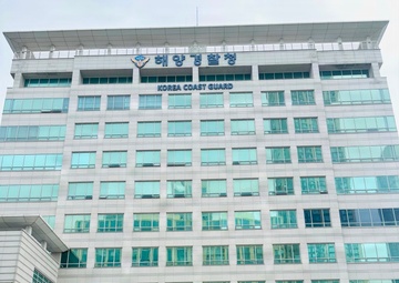 Four individuals are posing for a group photo in front of the Korea Coast Guard (해양경찰청) headquarters building. The group includes one person in U.S. Army multicam/OCP camouflage uniform, one civilian in a white shirt with a lanyard/badge, and two individuals in dark navy blue uniforms consistent with U.S. Coast Guard attire. The building is a large modern multi-story glass and stone structure with both Korean and English signage.